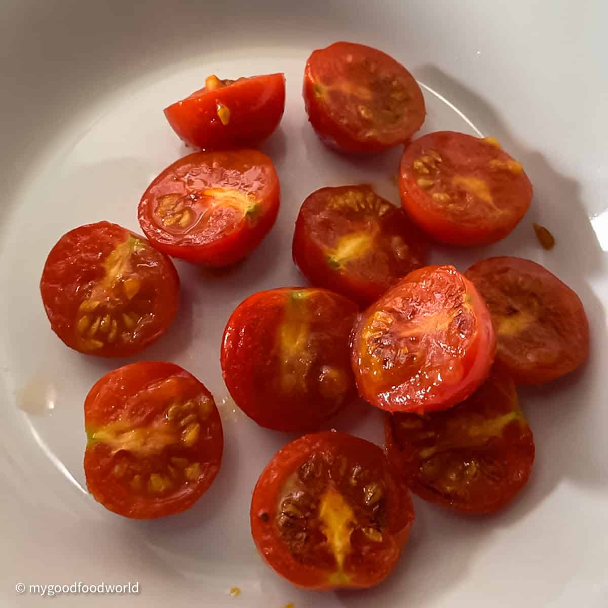 Cherry tomatoes halves that have been pan roasted in some olive oil are placed in a white bowl-plate.