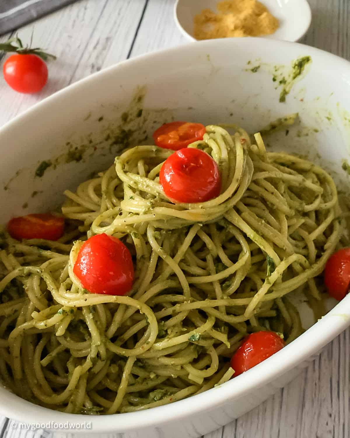 The dish pasta al pesto with pan roasted tomatoes is placed in a white oval dish. Next to the bowl, is placed one cherry tomato and some nutritional yeast in a small white colored bowl.