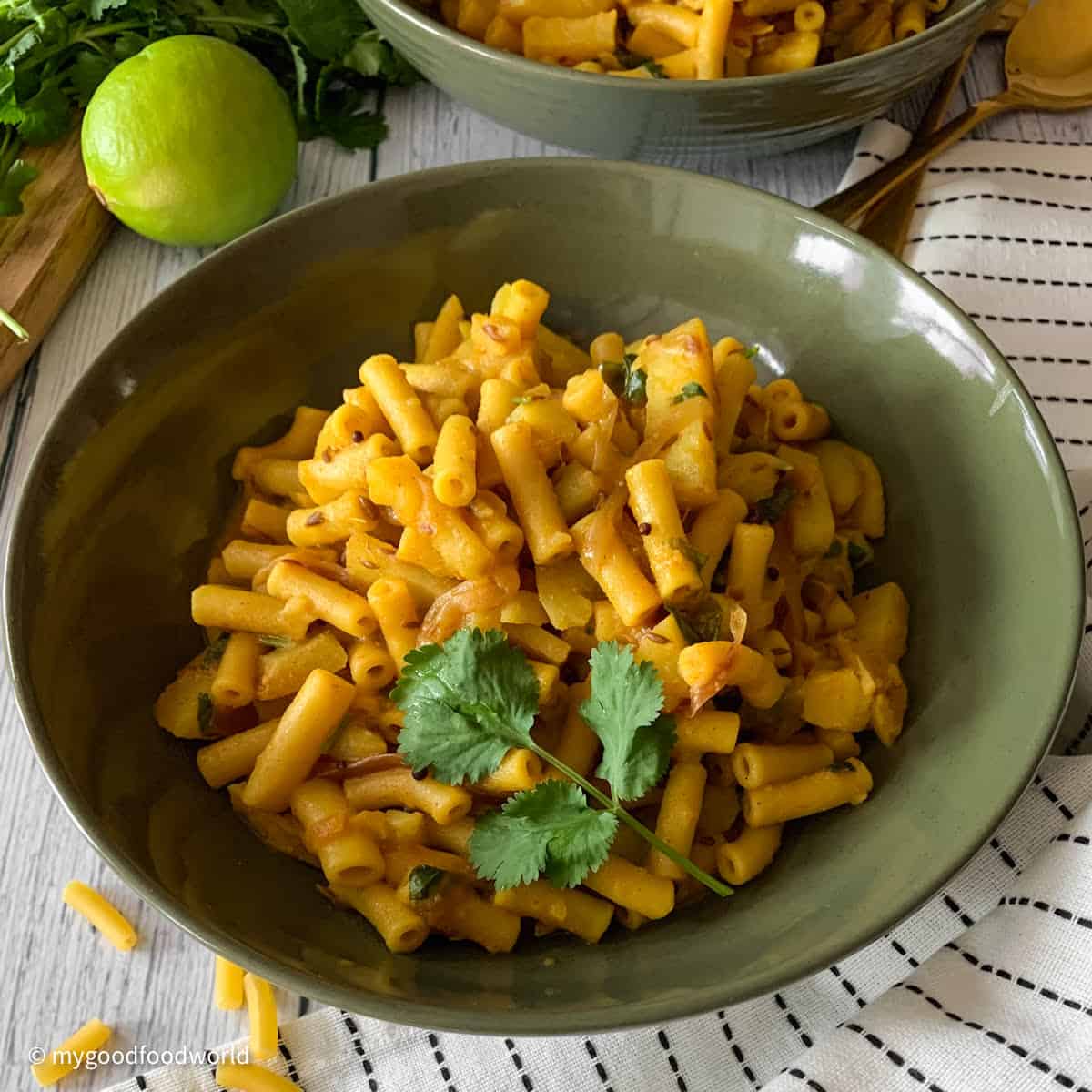 A deep yellow colored pasta dish that is garnished with a sprig of cilantro and spices such as cumin and mustard seeds is placed in a round bowl. The bowl is placed on a white napkin with black stripes.