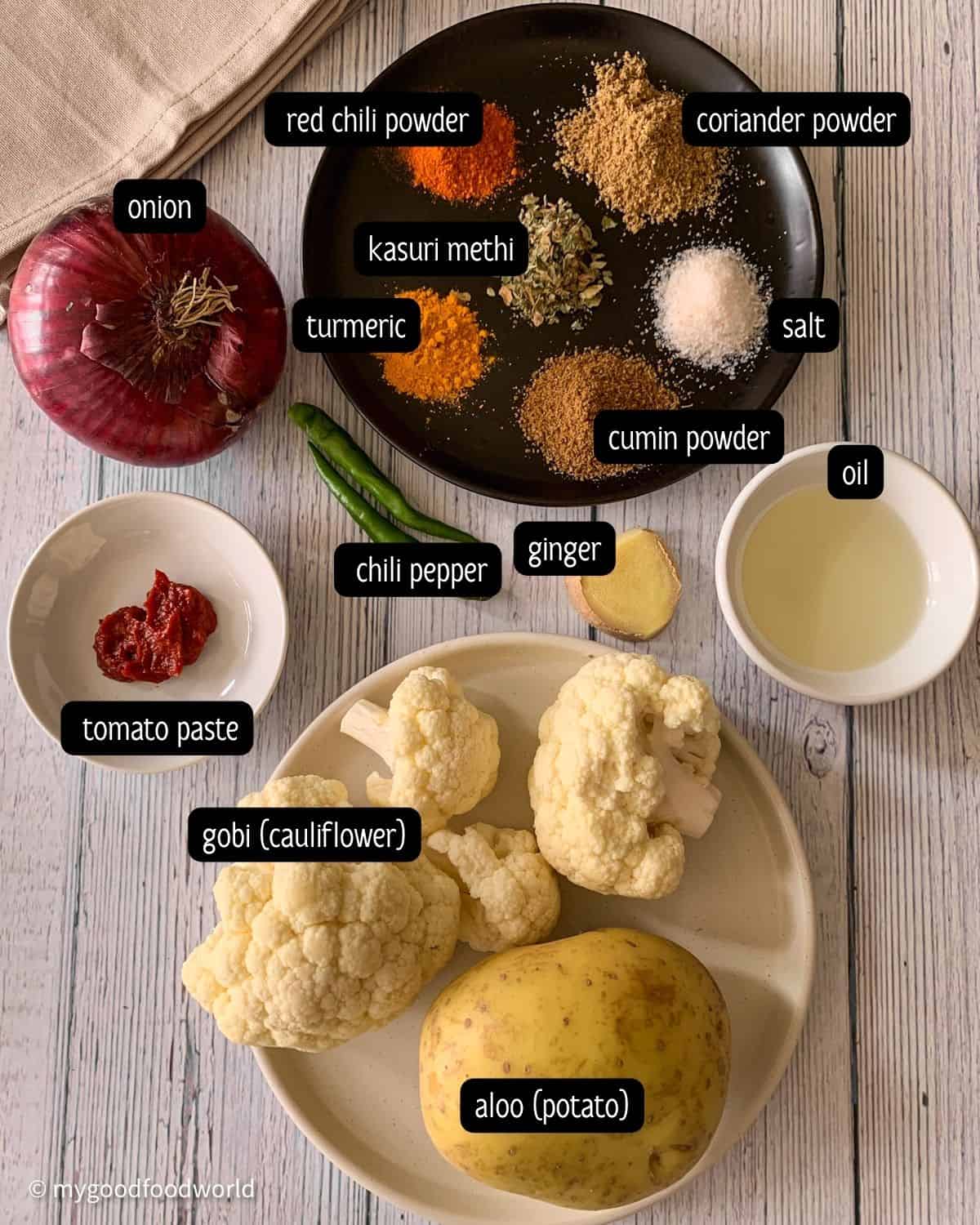 Various Indian spices such as red chili powder, kasuri methi, coriander powder and ingredients for aloo gobi sabzi are placed on a wooden table.