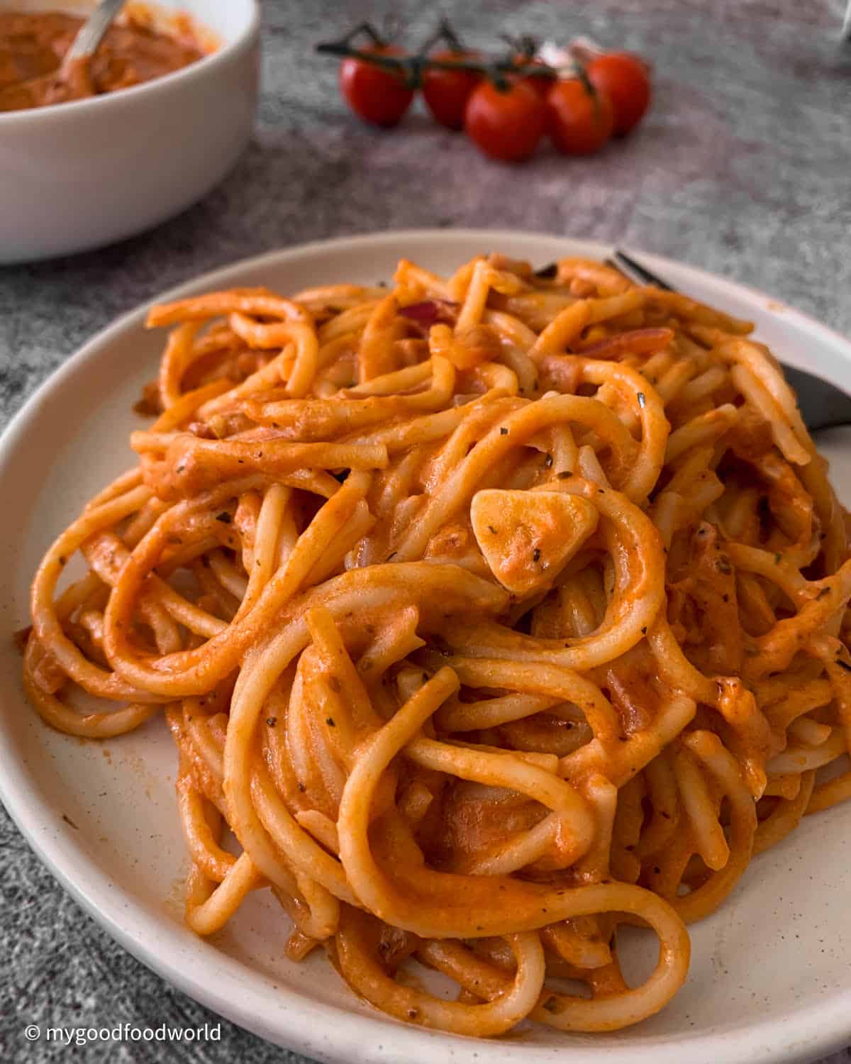 Some spaghetti coated with a creamy red sauce is placed in a heap on a white round plate. A slice of garlic is placed on the pasta. Some cherry tomatoes on the vine and a bowl of the sauce are also seen in the picture.