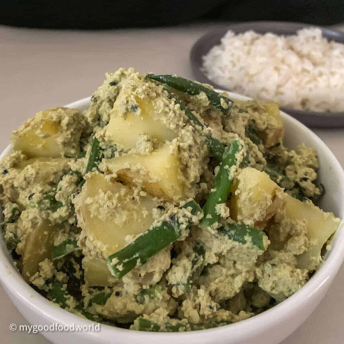 A bowl of green curry with aloo and green beans on a white plate with a side of rice.