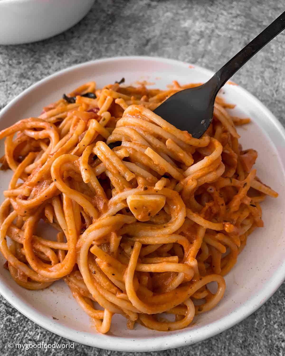 Creamy tomato sauce coated spaghetti pasta that has been served in a white round plate is being scooped with a black colored fork.