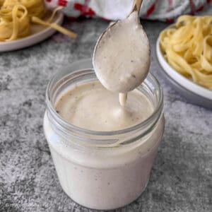 Creamy alfredo sauce is placed in a glass jar. Some of the sauce is dripping from a spoon into the jar.