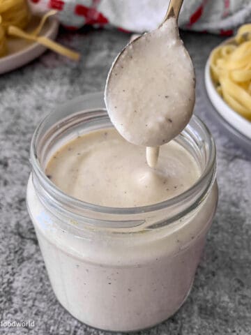 Creamy alfredo sauce is placed in a glass jar. Some of the sauce is dripping from a spoon into the jar.