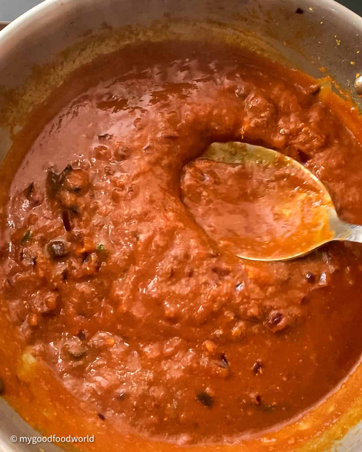 A close up photo of a pot of tomato curry gravy with a steel spoon in it.