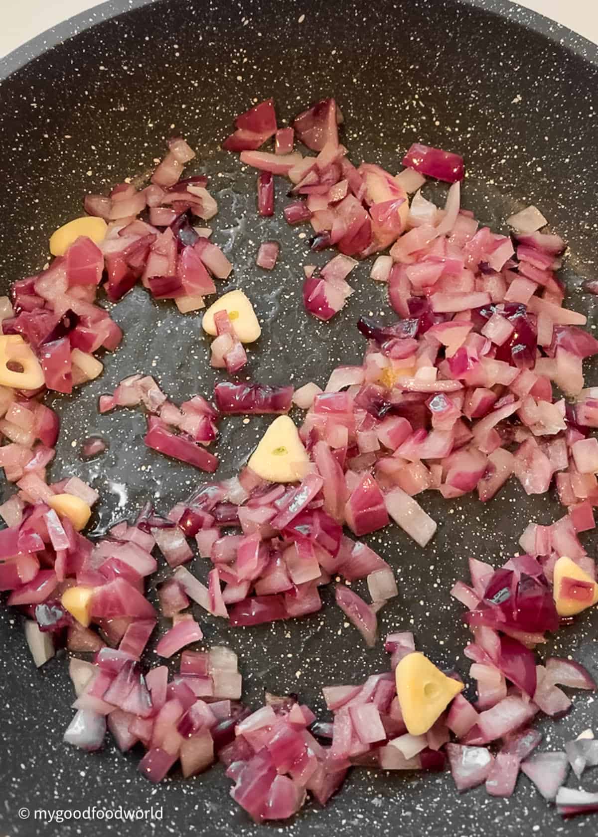 Finely chopped onions and garlic are frying in a dark grey skillet. The red onions are looking pale as they are frying.