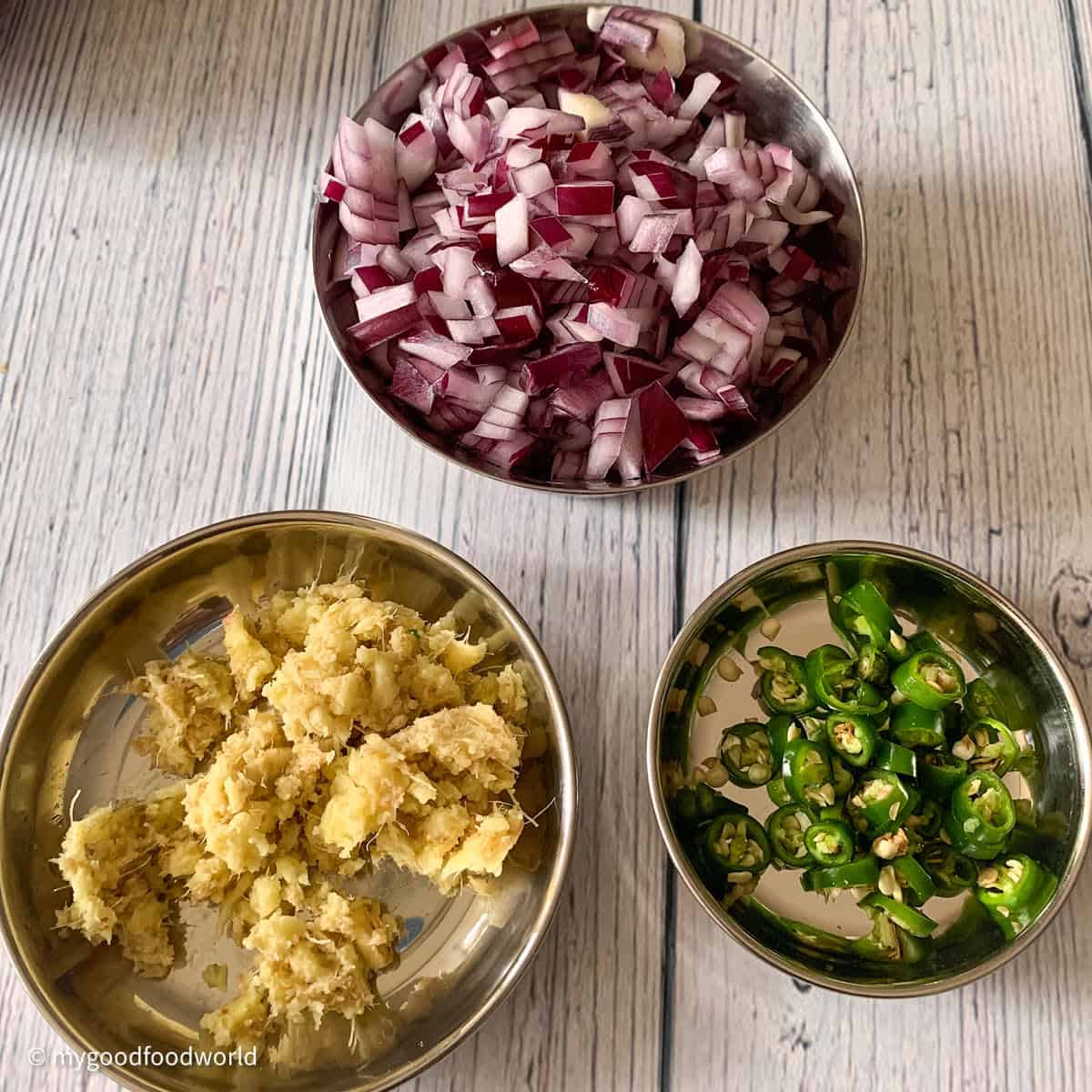 Three bowls of food ingredients, one with chopped red onions, one with crushed ginger, and one with sliced green chilies.