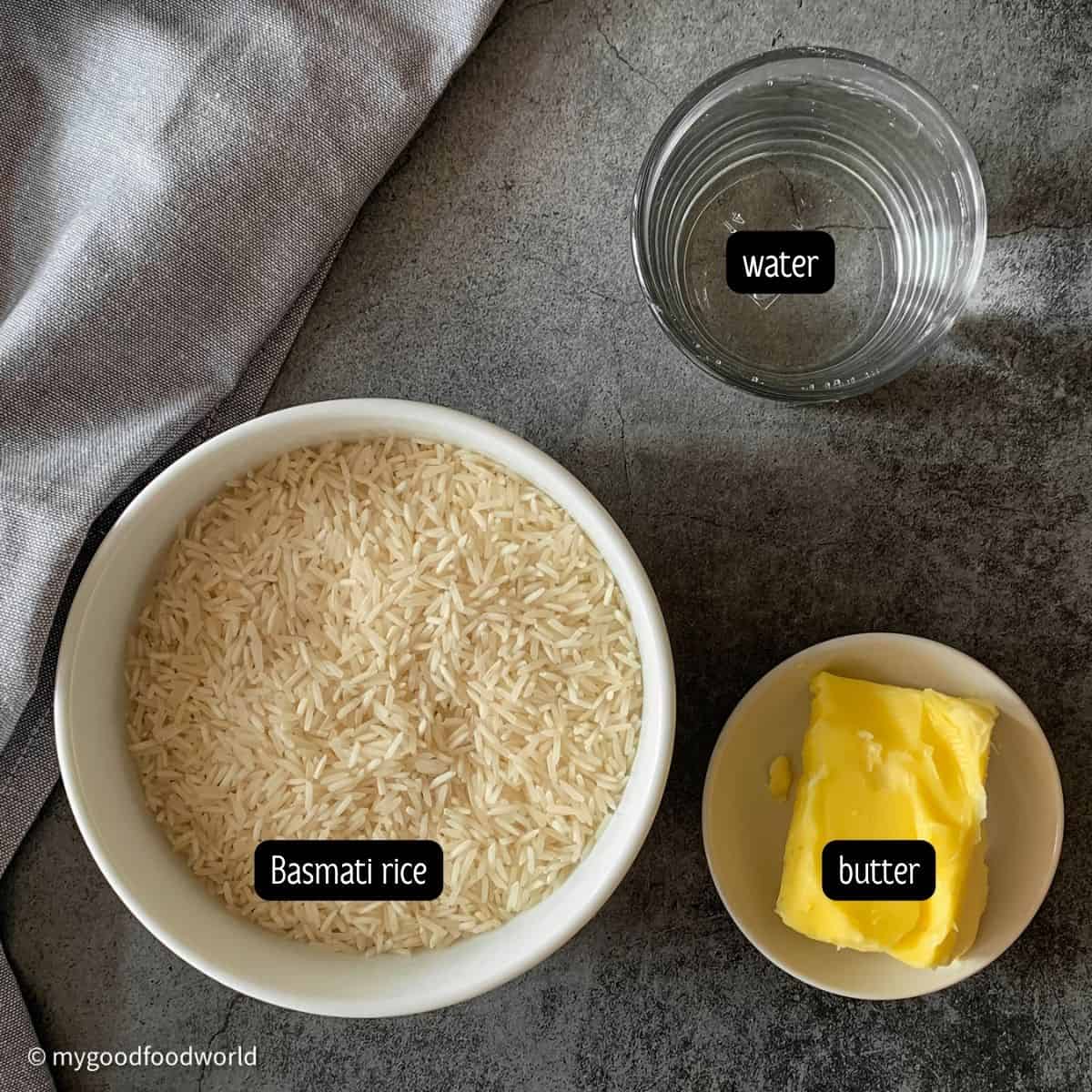 Some yellow butter, uncooked white rice, and some water are placed in bowls next to each other on a dark grey backdrop.