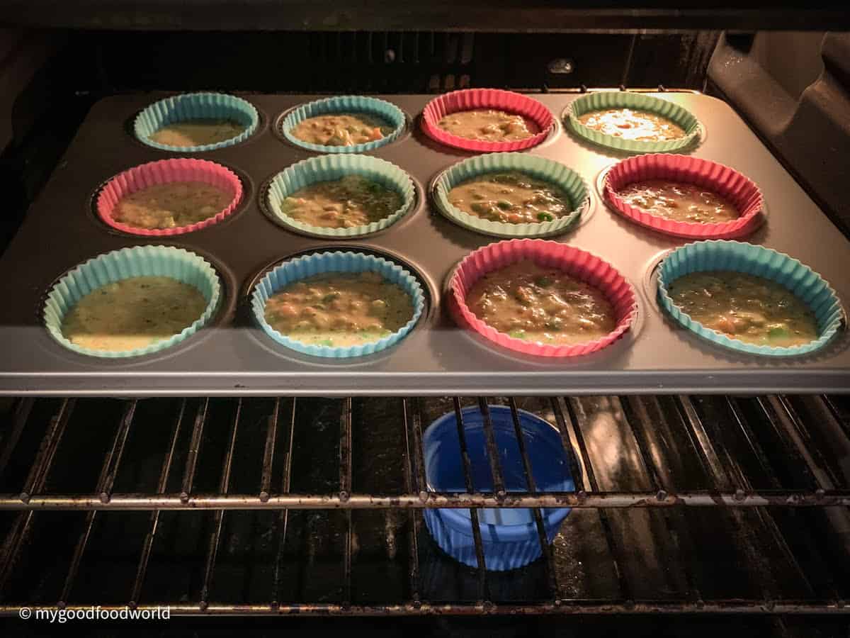 A baking tray with muffin batter-filled molds placed in the oven. A small blue colored bowl is placed on the floor of the oven.