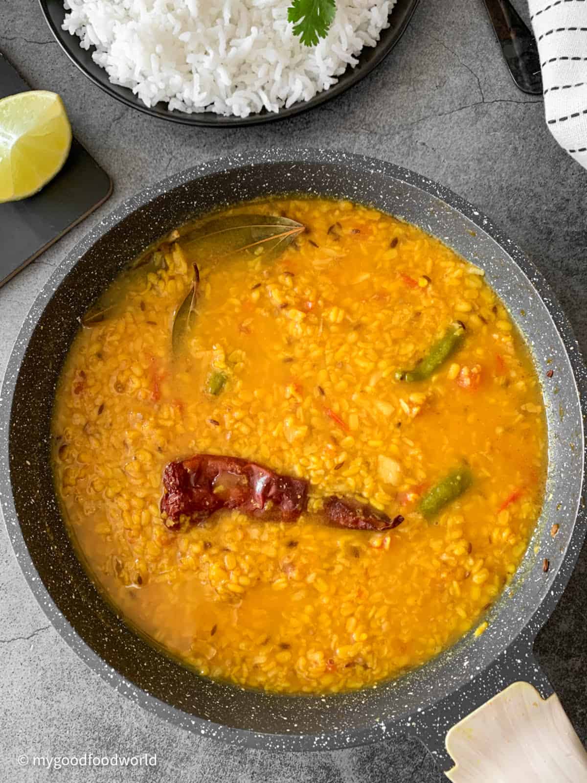Moong lentils cooked with simple spices such as cumin, tomatoes and chilies is placed in a skillet. The skillet is placed on a dark colored table where a piece of lime and some cooked white rice are also placed.