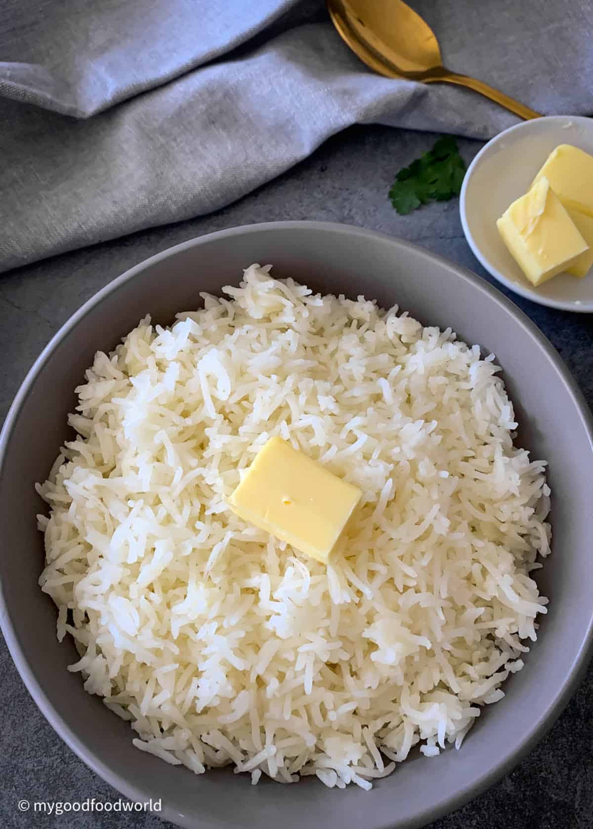 Cooked buttery rice is placed in a serving bowl with a cube of butter on top as garnish. Some more butter is placed in a plate next to the bowl, along with a gold color spoon, a cloth napkin and cilantro.