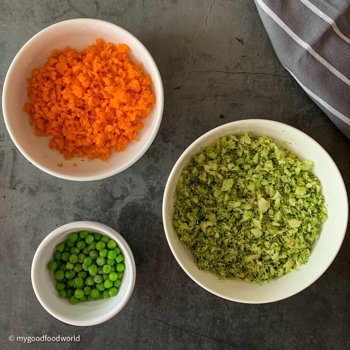 Finely chopped broccoli and carrots are placed in round white bowls along with a bowl of green peas.