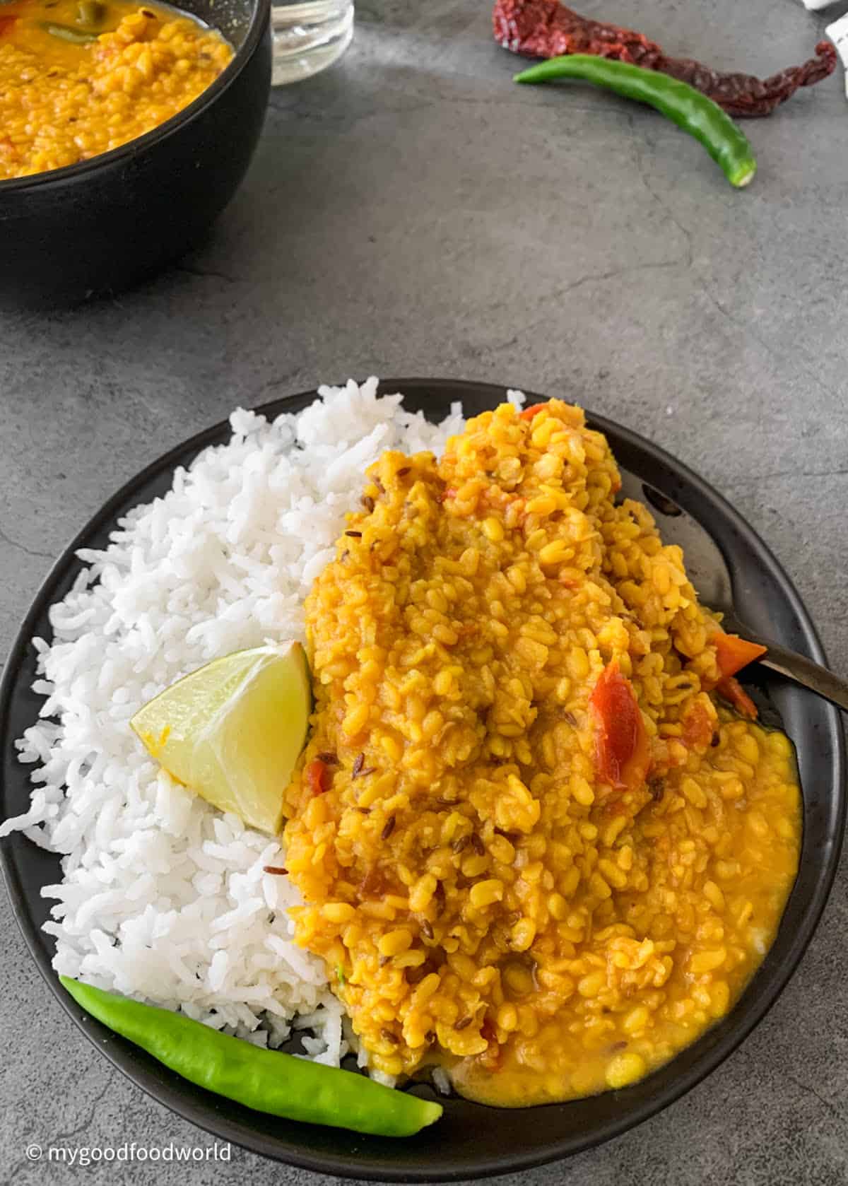 Bengali moong dal is served in a black plate with some cooked white rice, a chili pepper, and a piece of lime.