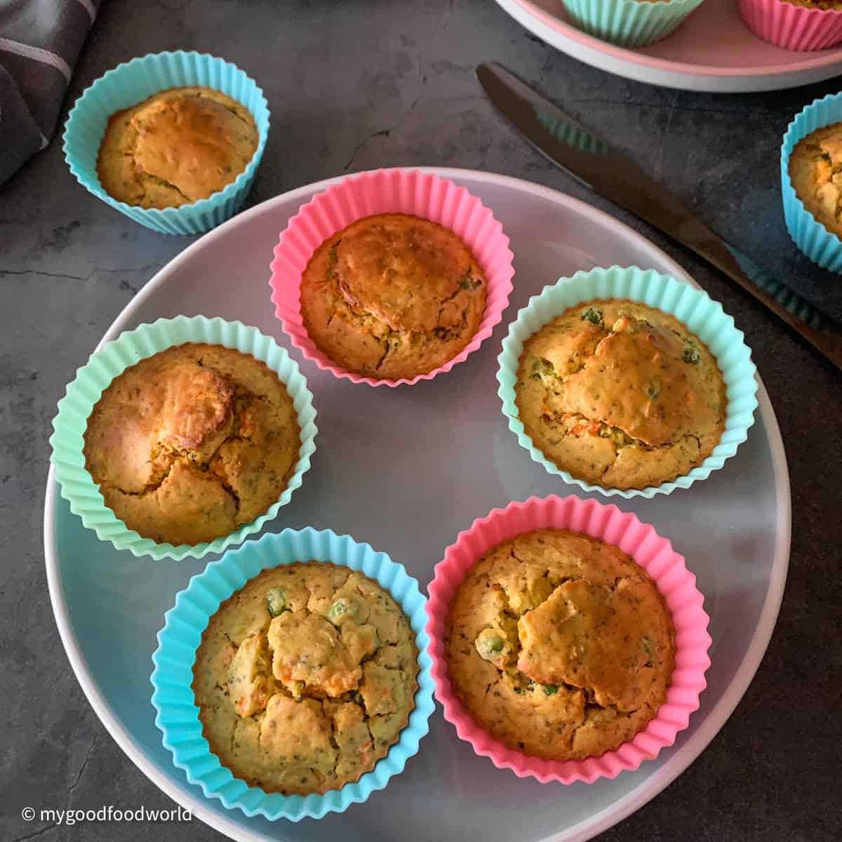 A plate of colorful silicone molds with baked muffins is placed on a dark grey background. Next the plate, a black colored butter knife and some more muffins in their molds are placed.