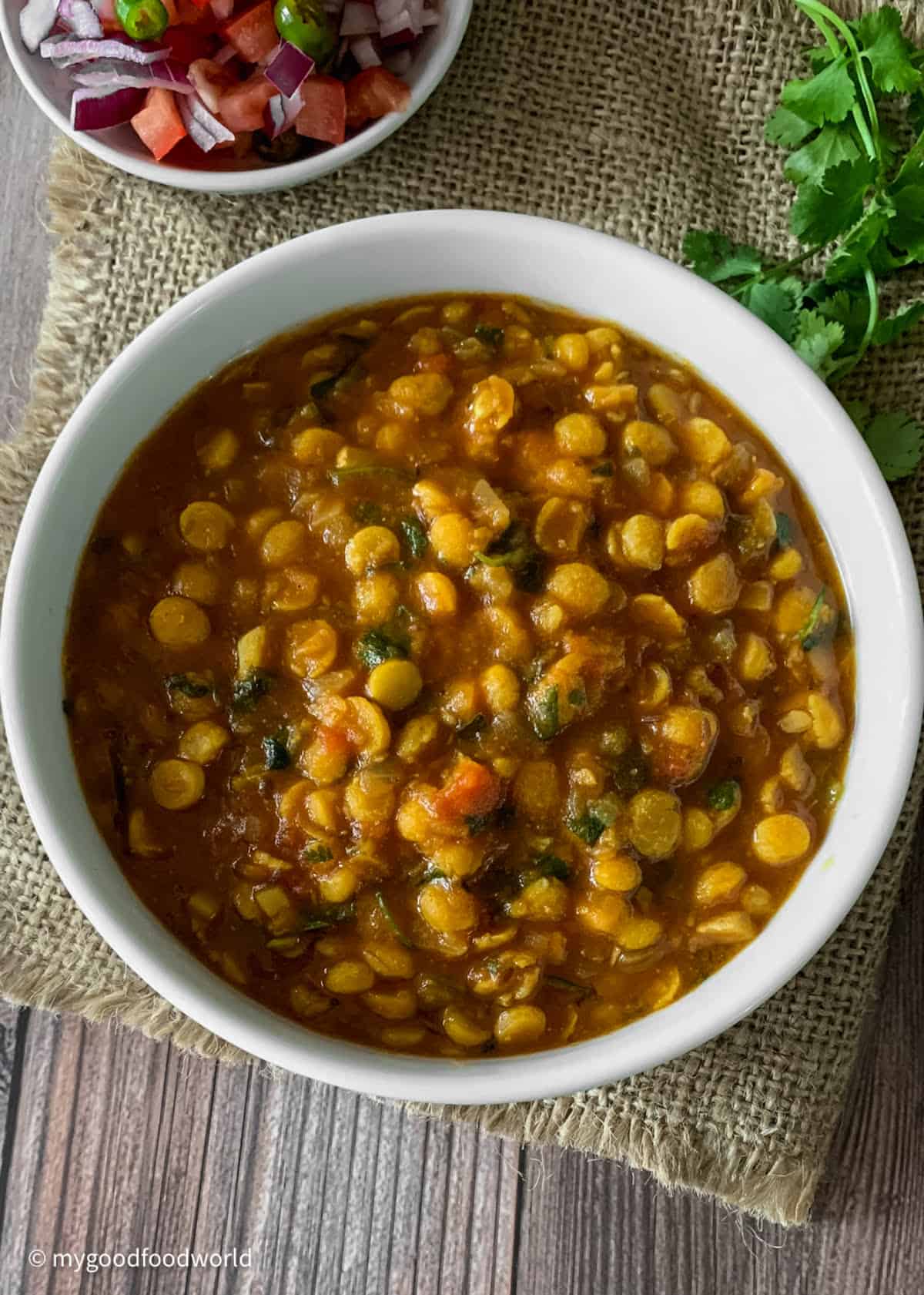 A bowl of chana dal cooked in a tomato sauce, topped with cilantro. The bowl is placed on a piece of burlap on a wooden table,