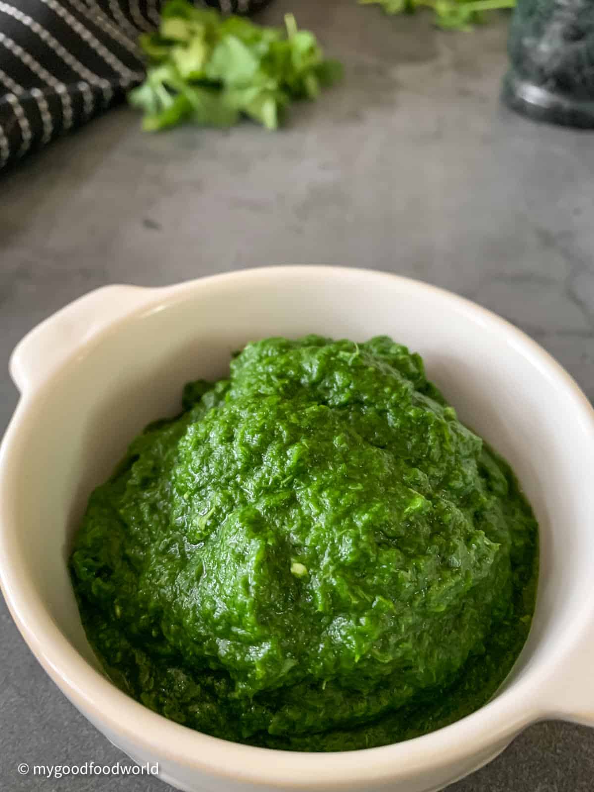 Green chutney is placed in a white bowl. Also seen are some fresh coriander leaves, a pestle and a cloth napkin.