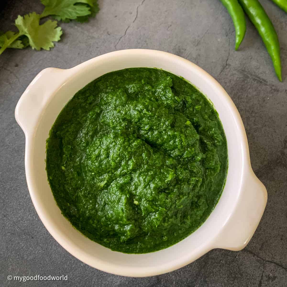 Freshly made Indian green chutney is placed in a white round bowl on a dark colored backdrop. Some fresh chilies and cilantro leaves are placed next to the bowl.