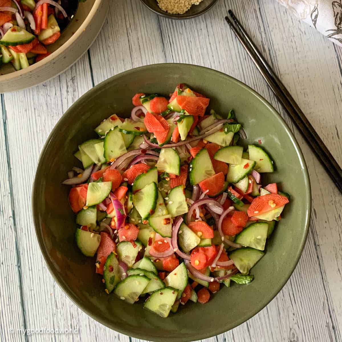 Salad of carrots and cucumber and red onions is served in a bowl. The salad has some toasted sesame seeds and red pepper flakes garnish.
