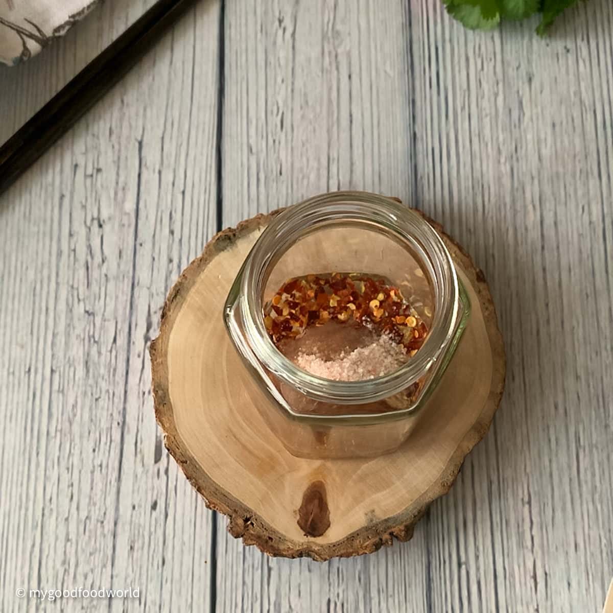 A glass jar with some red pepper flakes, salt and other ingredients are placed on a round wooden block.