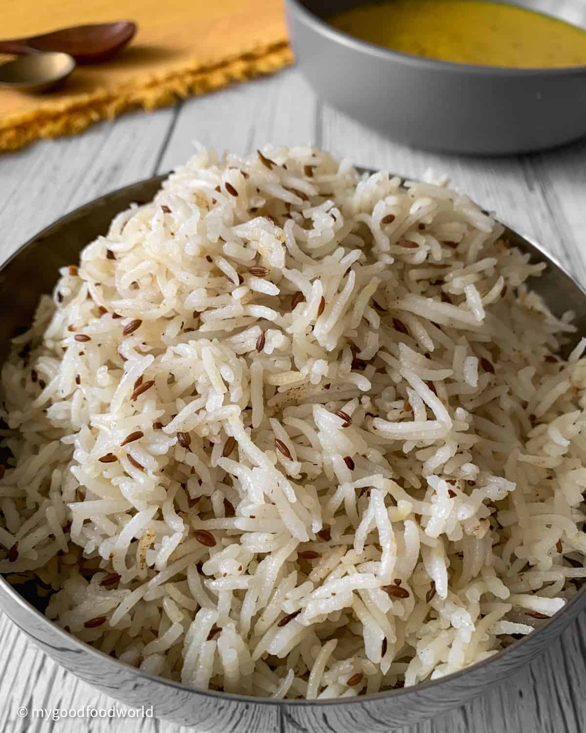 Cooked cumin rice is being served in a round stainless steel bowl on a white patterned background.