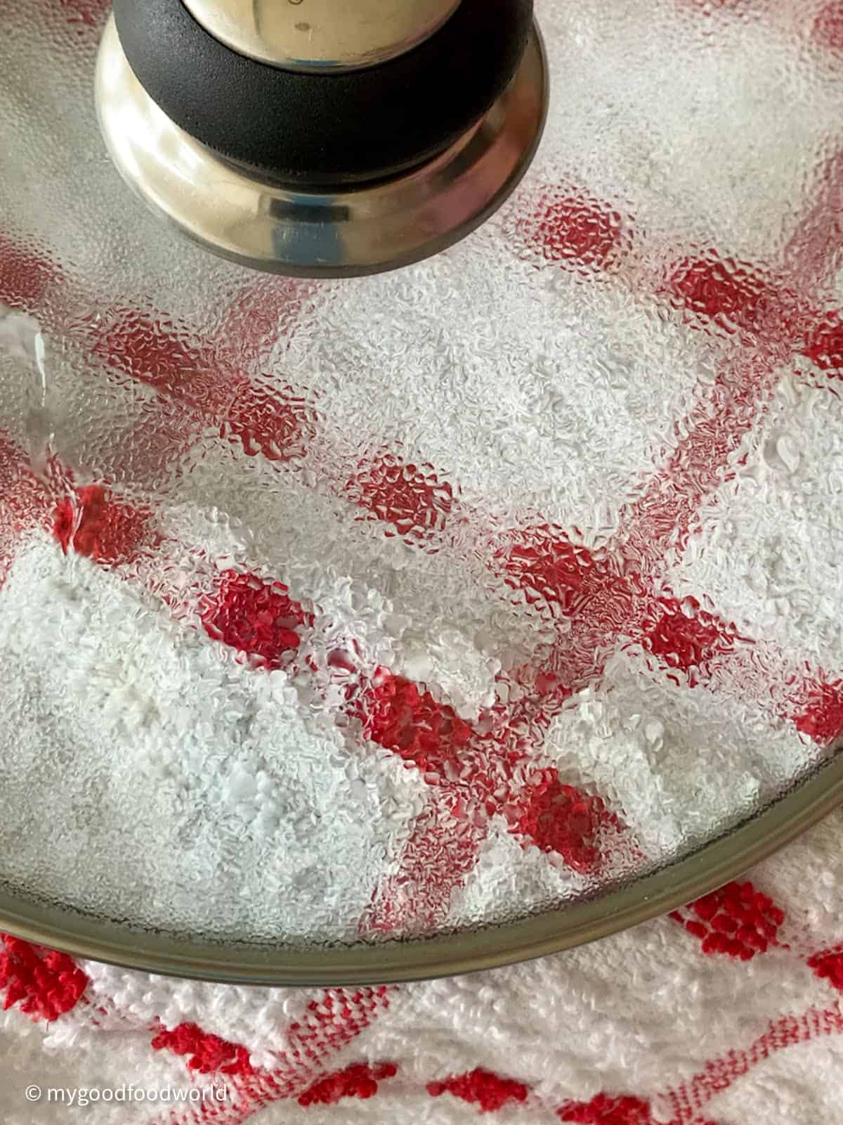 A red patterned white kitchen towel is covered by a round glass lid with a knob.