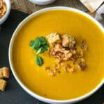 Healing sweet potato turmeric soup is served in a round white bowl. The soup is garnished with some fresh mint leaves and bread croutons.