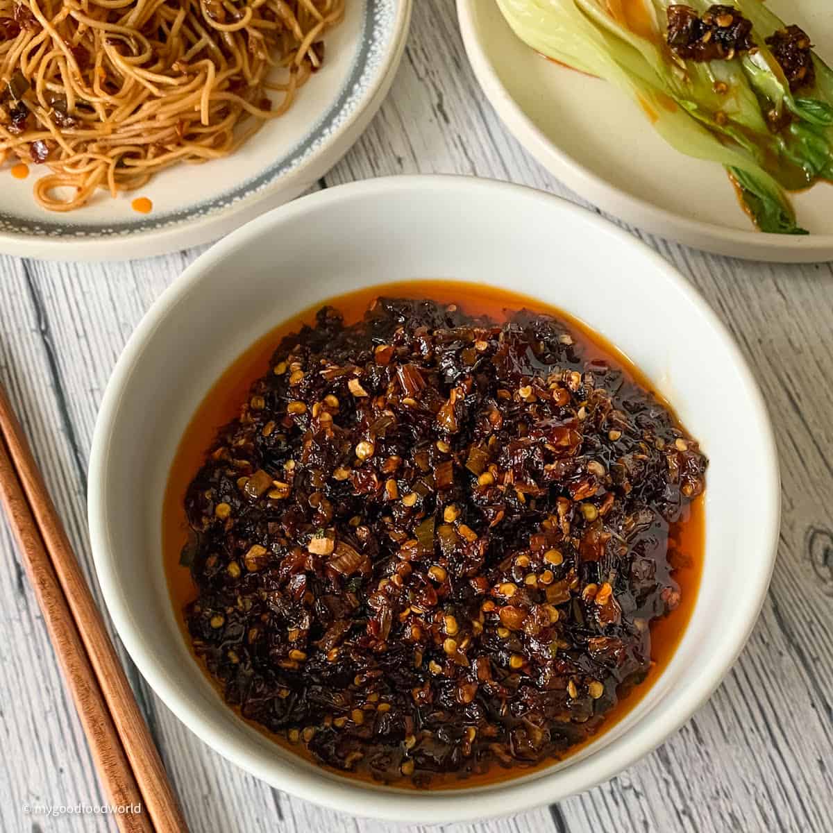 Chili oil made with herbs and spices is served in a bowl and placed on a table. Chopsticks and some noodles are placed next to the bowl.