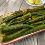 Whole tender okra that have been sauteed in powdered spices are placed in a neat arrangement in a rectangle-shaped plate with some wedges of lemon on the side.
