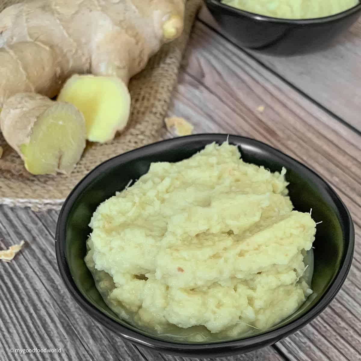 Fresh ginger puree is placed in a round black bowl on a brown tabletop. Next to the bowl, on a piece of burlap, is placed a piece of root ginger.