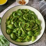 Pasta and spinach sauce are served in a round white lunch bowl. The bowl is placed on a grey colored table.