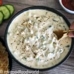 A bowlful of creamy cucumber raita is served in a round bowl. A hand with roti is scooping the raita.