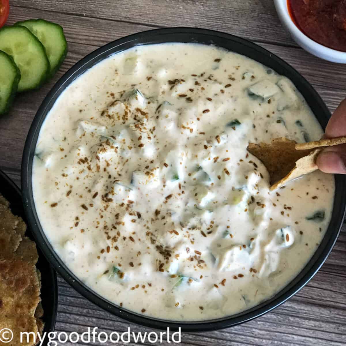A bowlful of creamy cucumber raita is served in a round bowl. A hand with roti is scooping the raita.