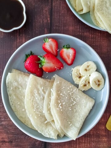 Three dairy-free coconut milk crepes are placed on a plate with some sliced bananas and strawberries.