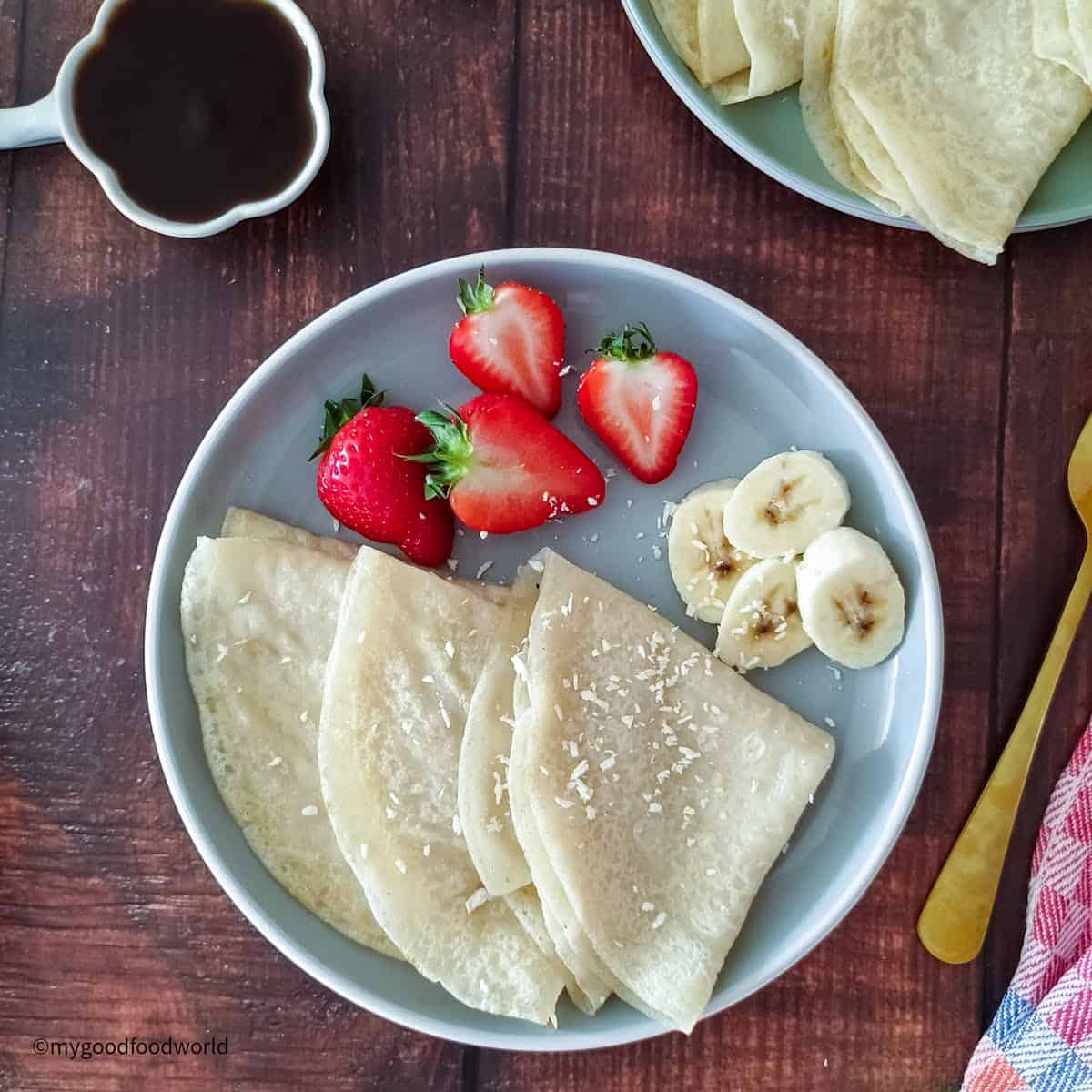 Three dairy-free coconut milk crepes are placed on a plate with some sliced bananas and strawberries.