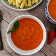 Creamy pasta sauce made with red peppers is served in a bowl. Next to the bowl is some cooked pasta on a plate.