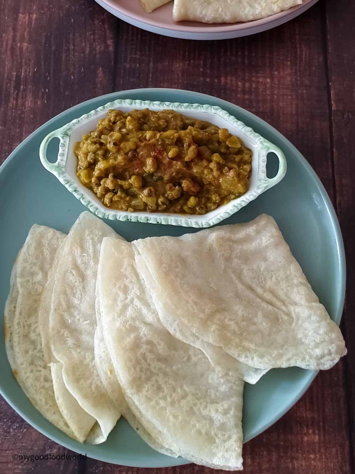 Three crepes made with coconut milk are folded and placed in a placed with a bowl of curry next to them.
