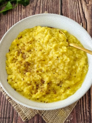 A simple kitchari recipe, made with mung lentils, rice, and spices such as ginger and black pepper, is served in a white bowl with a golden spoon.