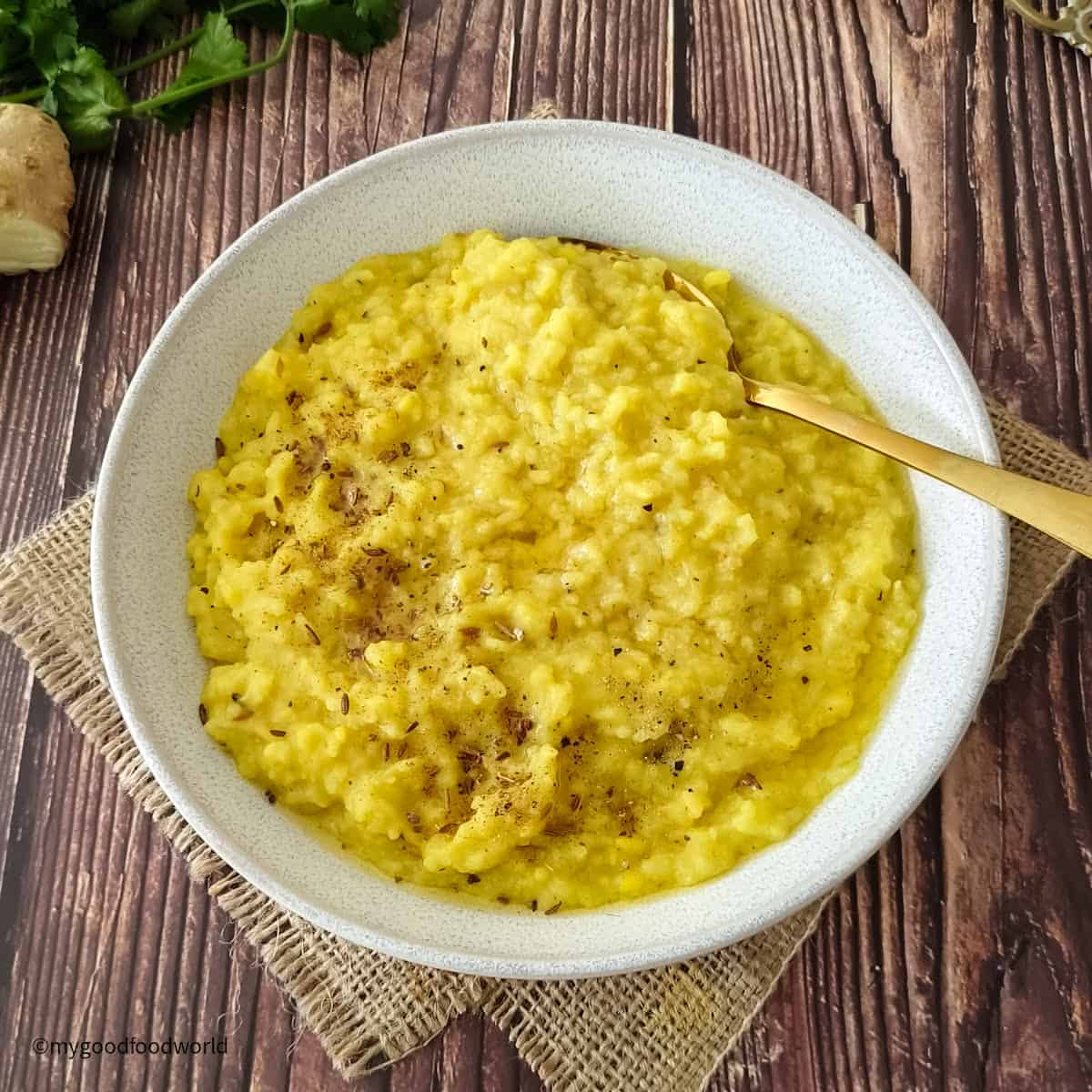 A simple kitchari recipe, made with mung lentils, rice, and spices such as ginger and black pepper, is served in a white bowl with a golden spoon.