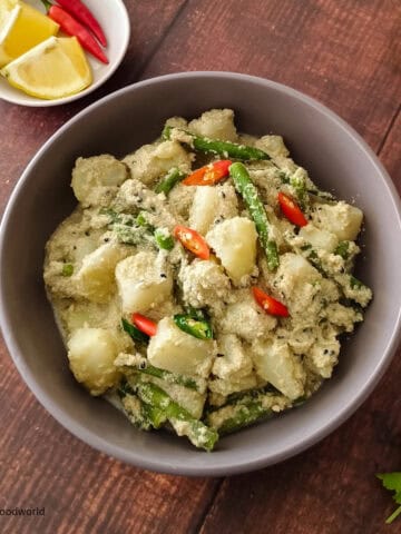 Bengali-style aloo posto is served in a round bowl with chopped chili pepper garnish. Some wedges of lemon and cilantro are placed next to the bowl.