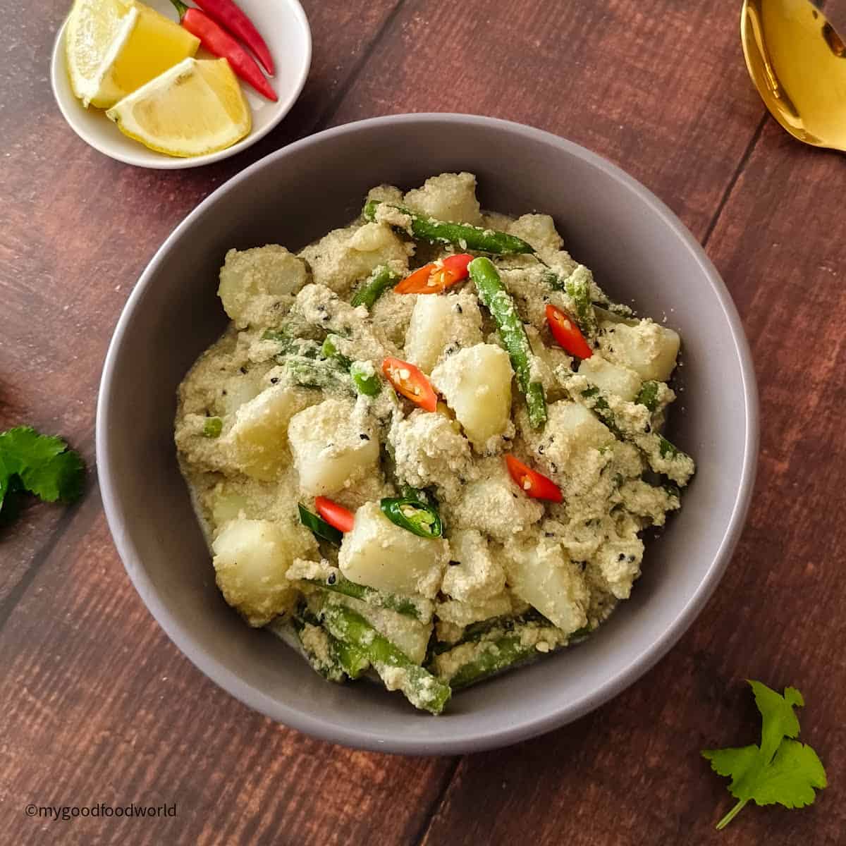 Bengali-style aloo posto is served in a round bowl with chopped chili pepper garnish. Some wedges of lemon and cilantro are placed next to the bowl.
