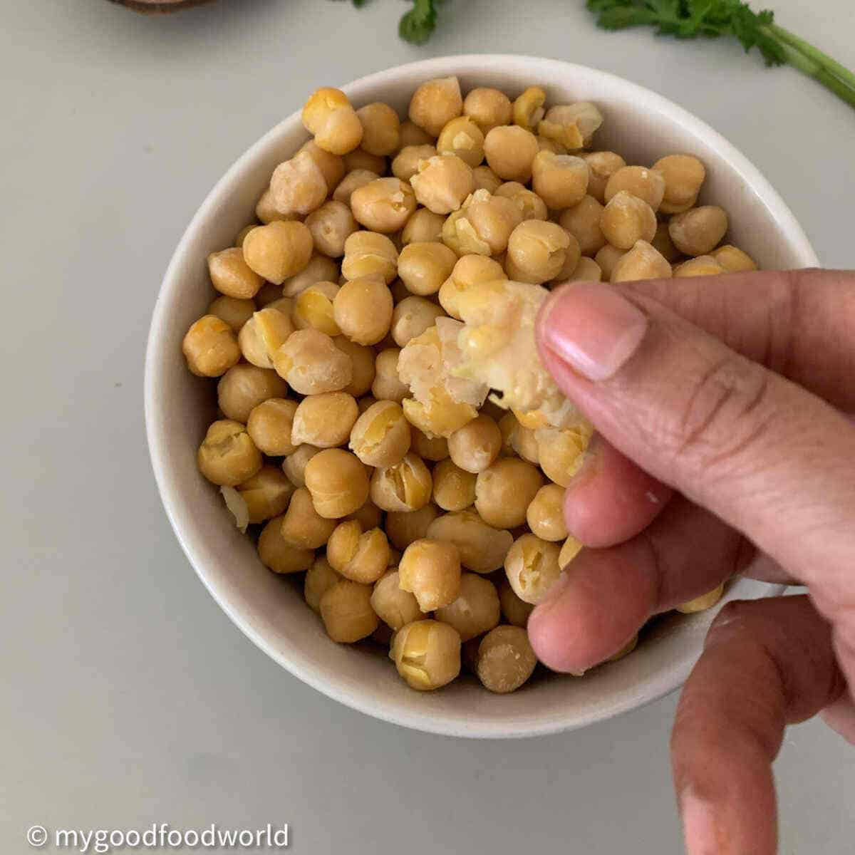 Perfectly cooked, soft and tender chickpeas are placed in a bowl. One chickpea has been squished between two fingers.