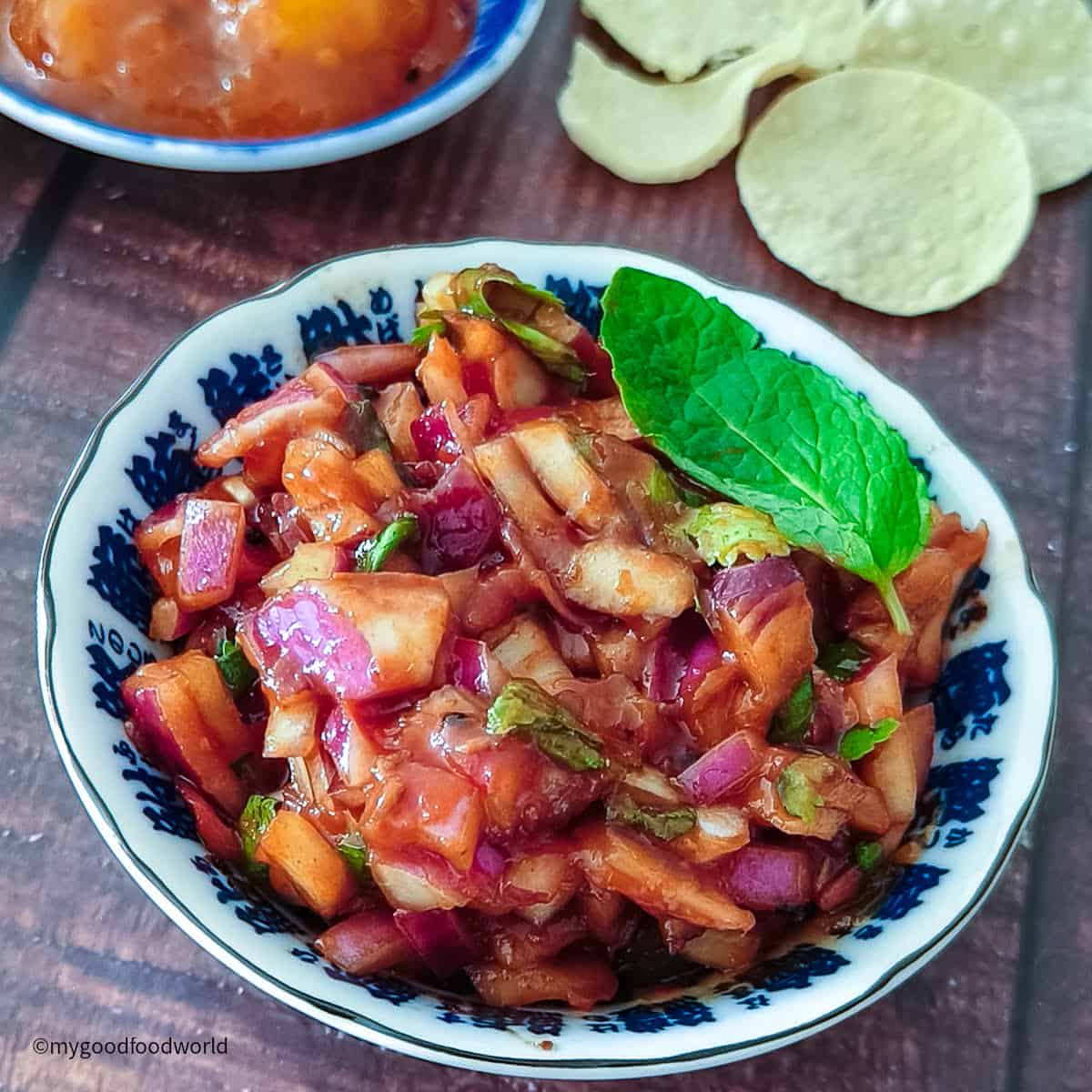 Close up of red spiced onion salad in a blue bowl served with poppadoms and mango chutney.