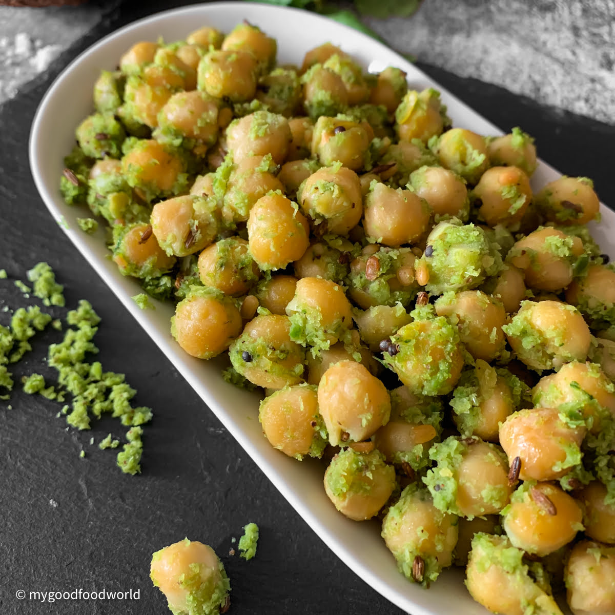 A close up shot of white chickpeas that have been tossed in a green color spice mix with whole cumin and mustard seeds.