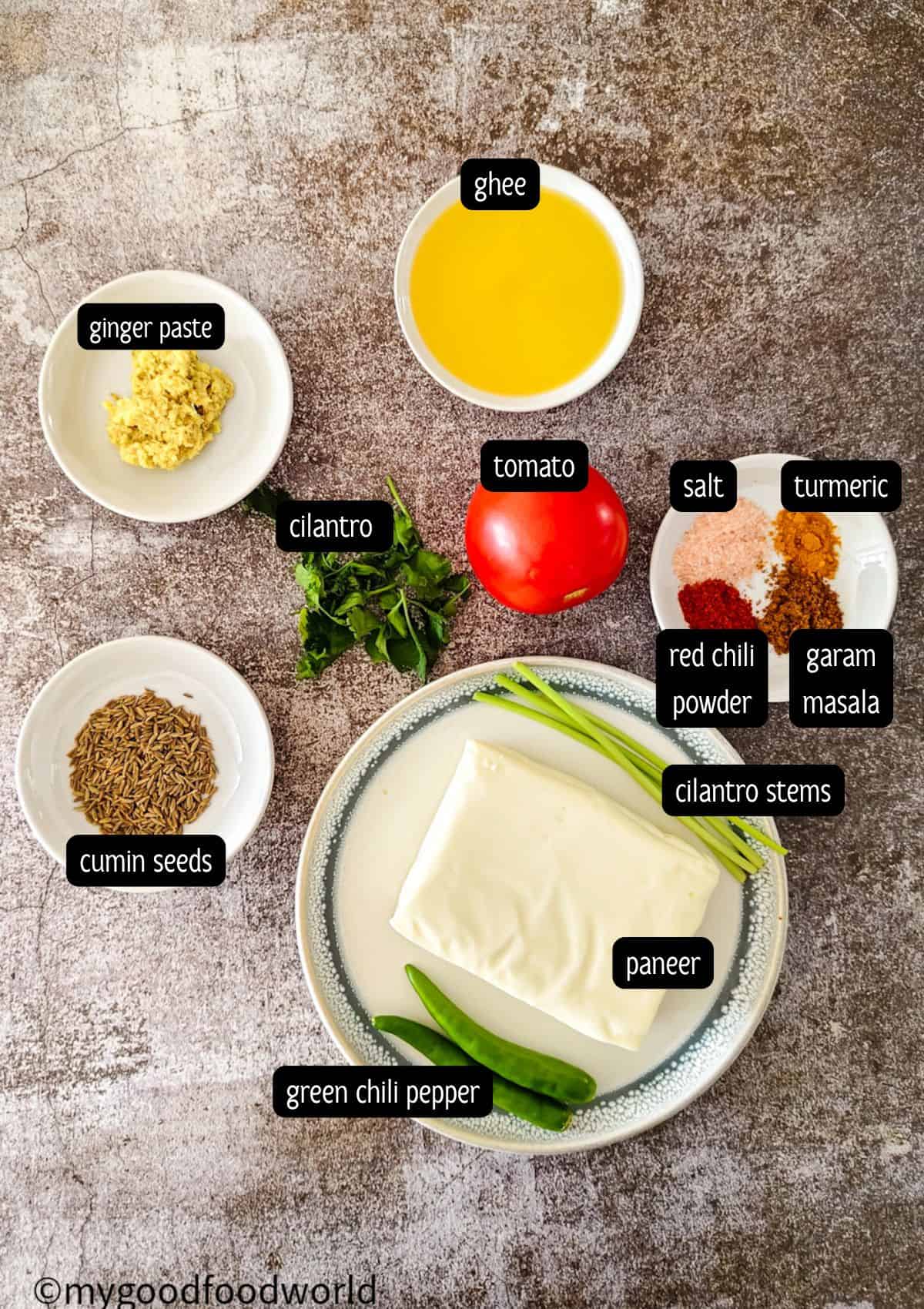 Overhead shot of ingredients for paneer bhurji including a block of paneer, tomato, green chilies, cumin seeds, coriander leaves, ghee, and mixed spices in a plate.