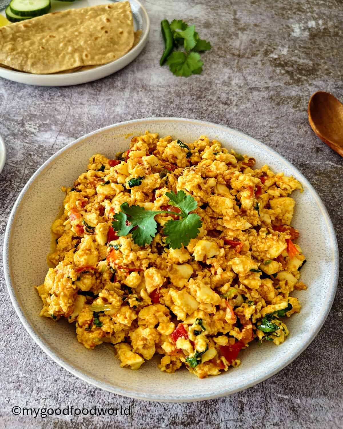 Angled view of a bowl of paneer bhurji next to a plate of roti, cucumber, and lemon wedges on a rustic stone background.