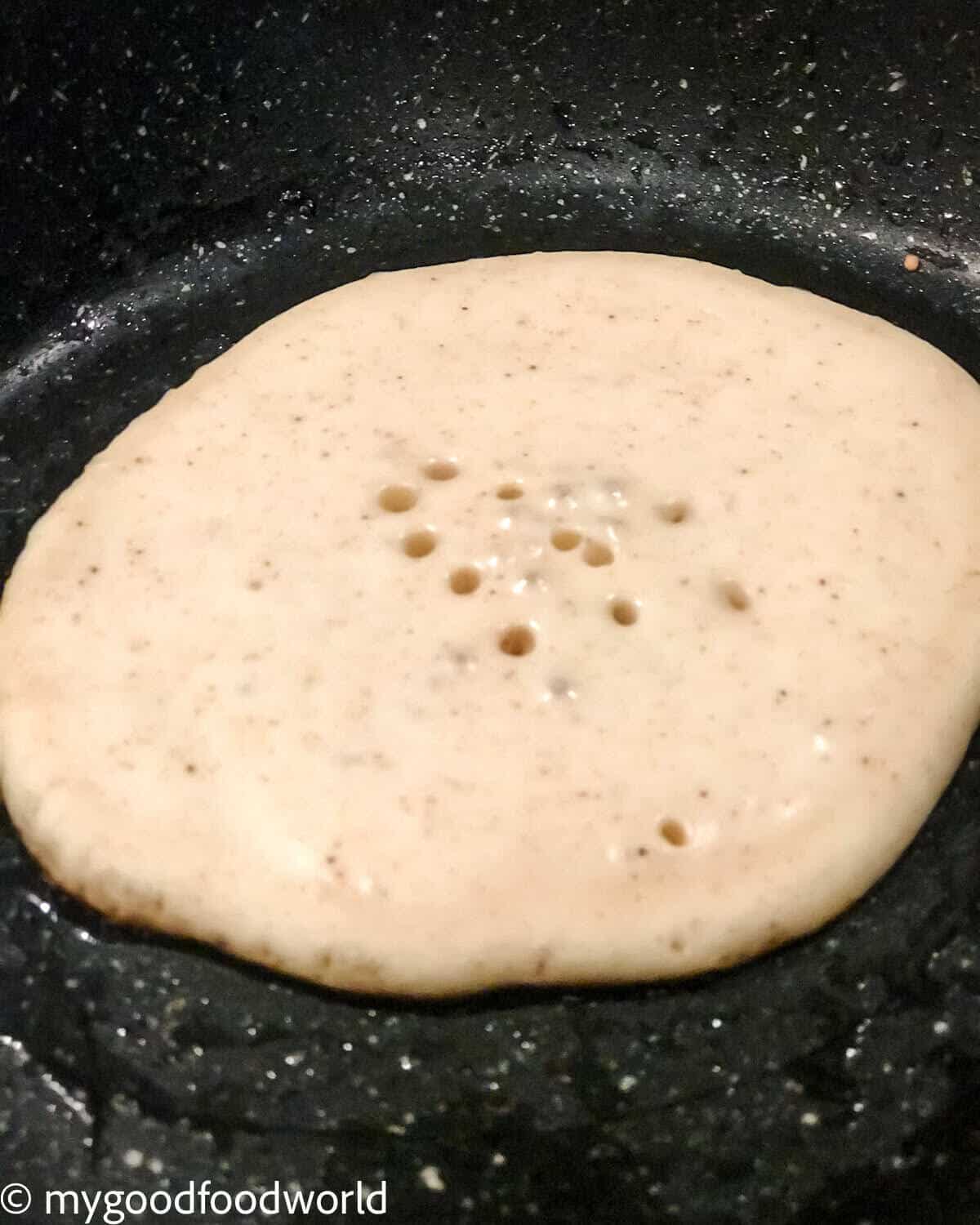 A pancake without eggs cooking in a skillet, with air bubbles forming on top before the first flip.