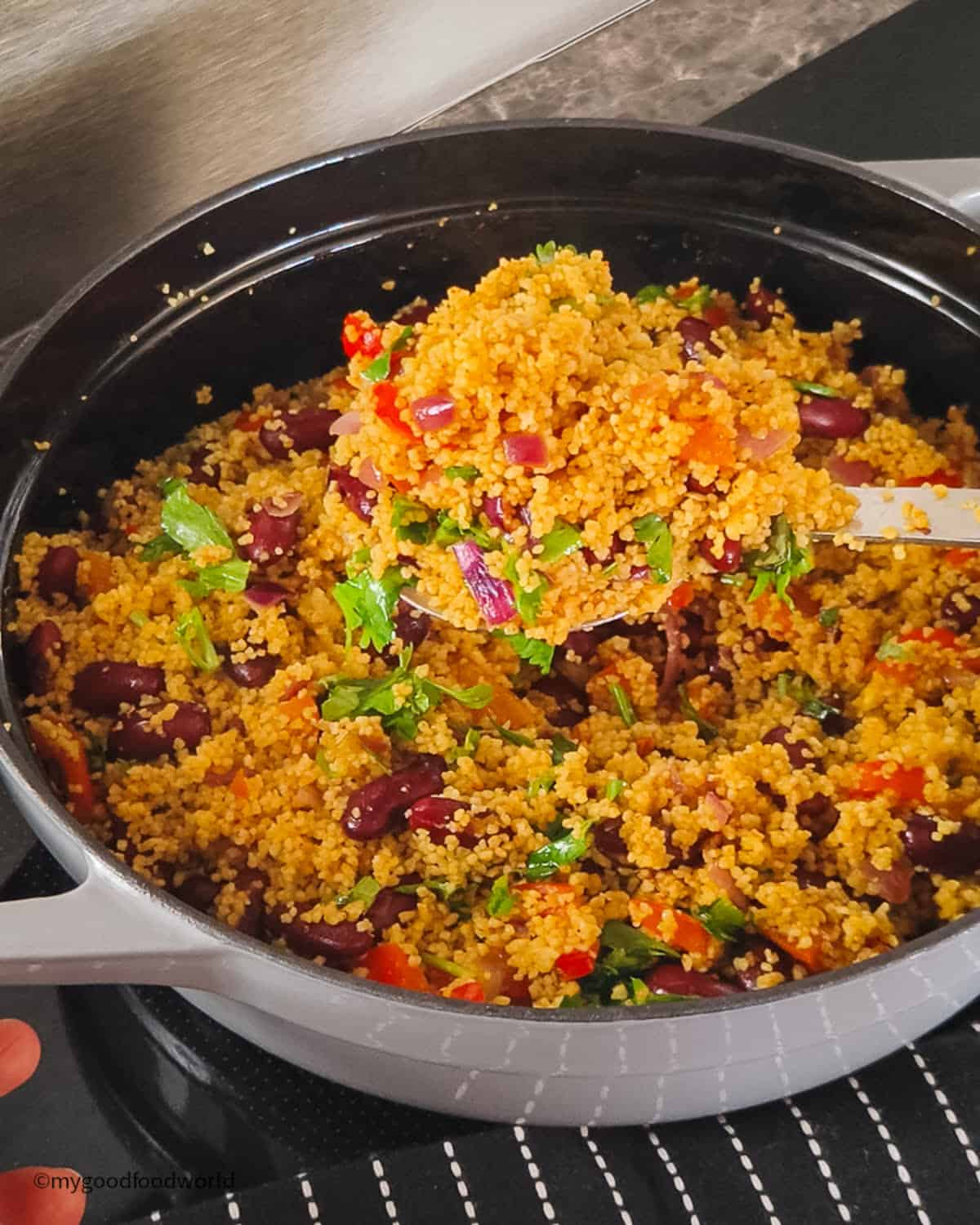 A spoon lifts a scoop of spicy couscous salad with kidney beans, peppers, and cilantro from the pan.