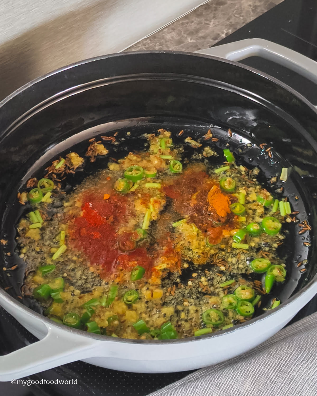Chopped chili peppers, cilantro stems, ginger paste, and spices such as red chili powder are being sauteed in a pan.