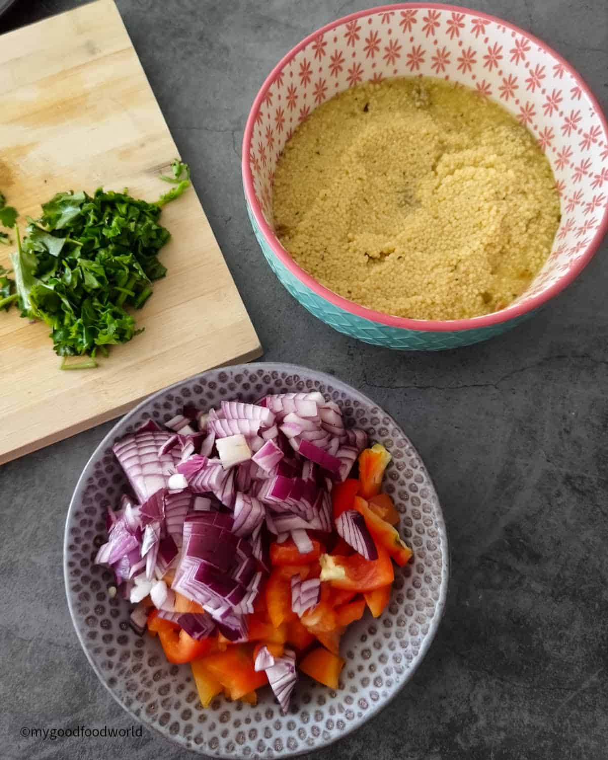 Couscous soaking in stock next to a bowl of chopped red onion and red bell pepper, with cilantro on a wooden board.
